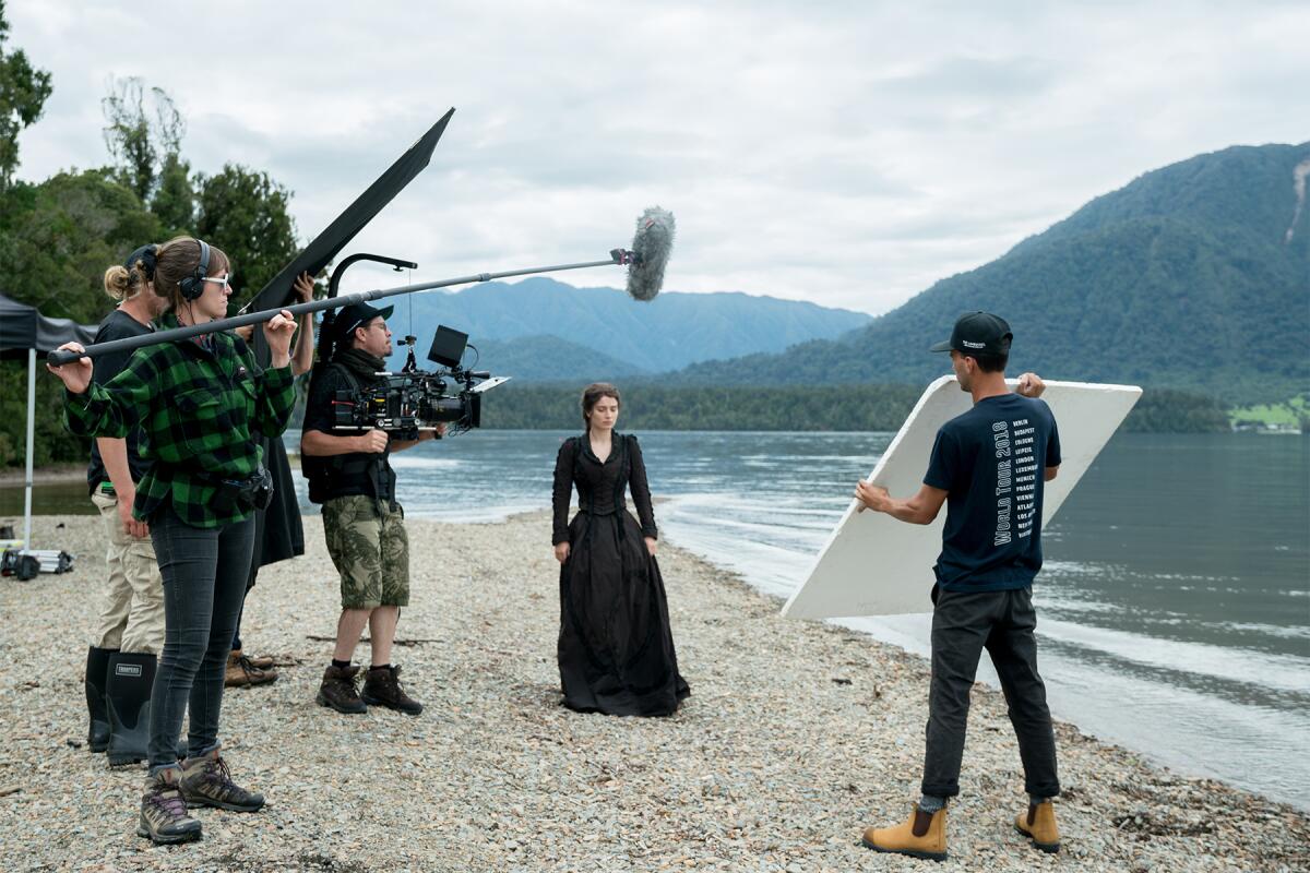 A woman in a black dress by the water, surrounded by a film crew.