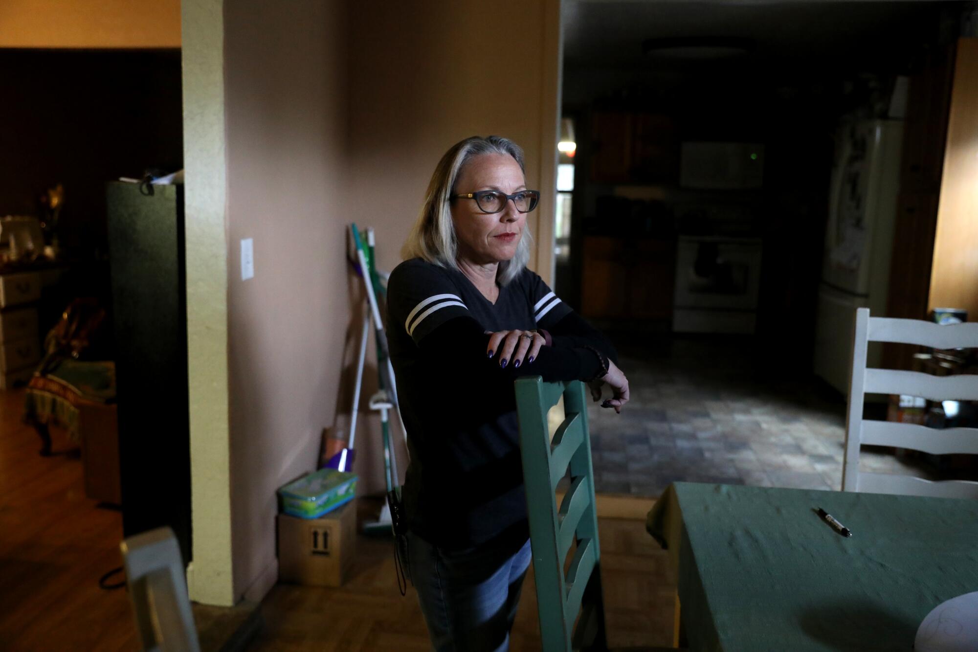 A woman stands inside a home's kitchen, leaning against a ladderback chair.