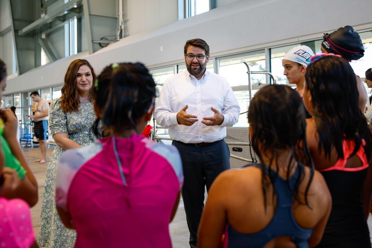 A woman in a dress, left, and a man  in white shirt and pants speak to a group of young people in athletic clothing