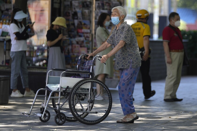 An elderly woman wearing a mask pushes her wheelchair past residents lined up for coronavirus testing, Thursday, July 7, 2022, in Beijing. (AP Photo/Ng Han Guan)