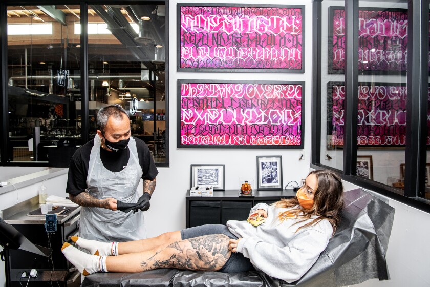 A man puts on gloves as a woman with tattoos on her leg lies on a reclined chair in a tattoo studio