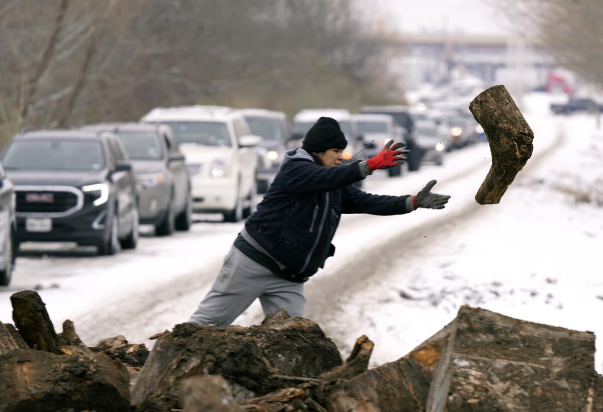 A woman tosses a log of firewood, with a long line of cars on a snowy road in the background