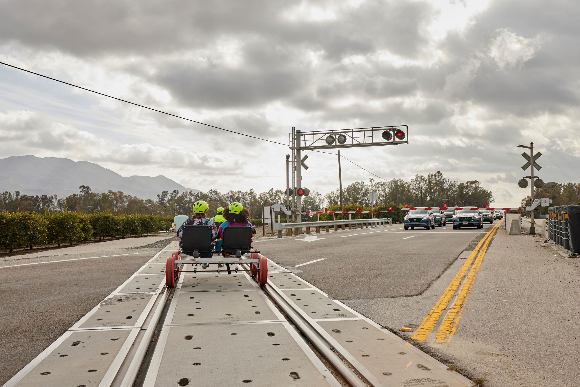 A view from behind of two people in a railbike, crossing a road with cars waiting for them to pass.