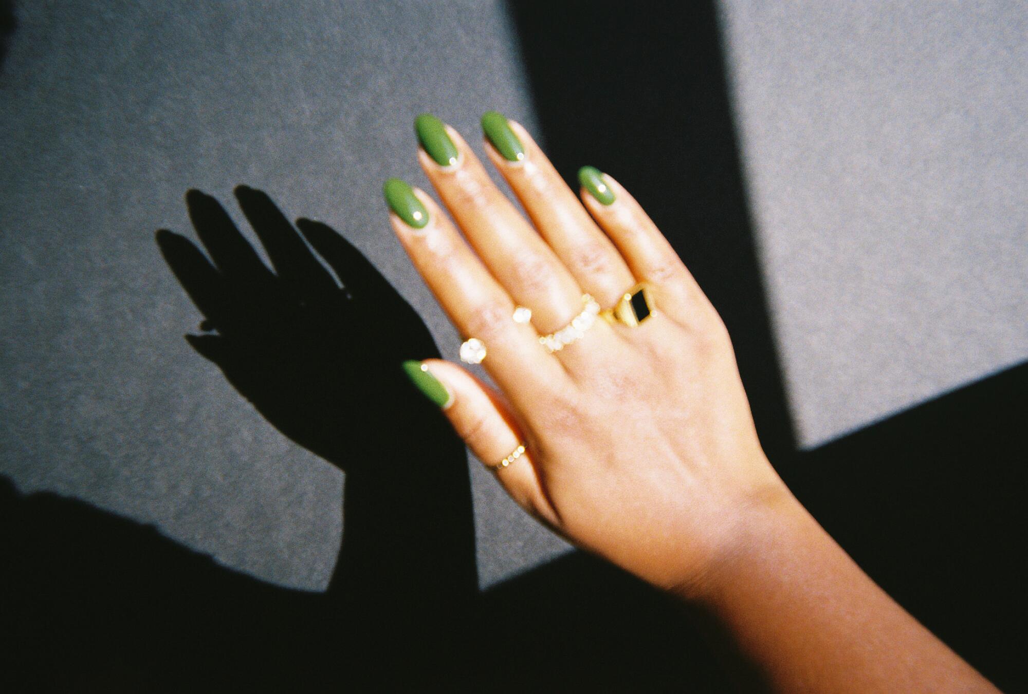 a soft-focus photo of a hand with green colored nails and various rings raised in front of a gray wall causing a harsh shadow