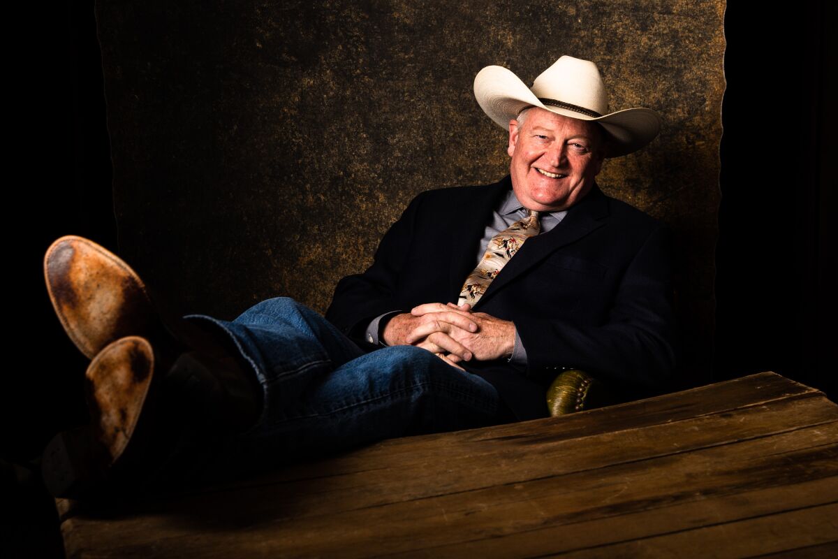 A man in a cowboy hat, jeans and jacket with tie smiles as he leans back in a chair with his feet propped on a table.