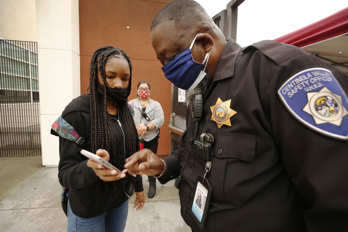 A school safety officer in a uniform with a badge views the screen of a student's phone. Both wear masks.