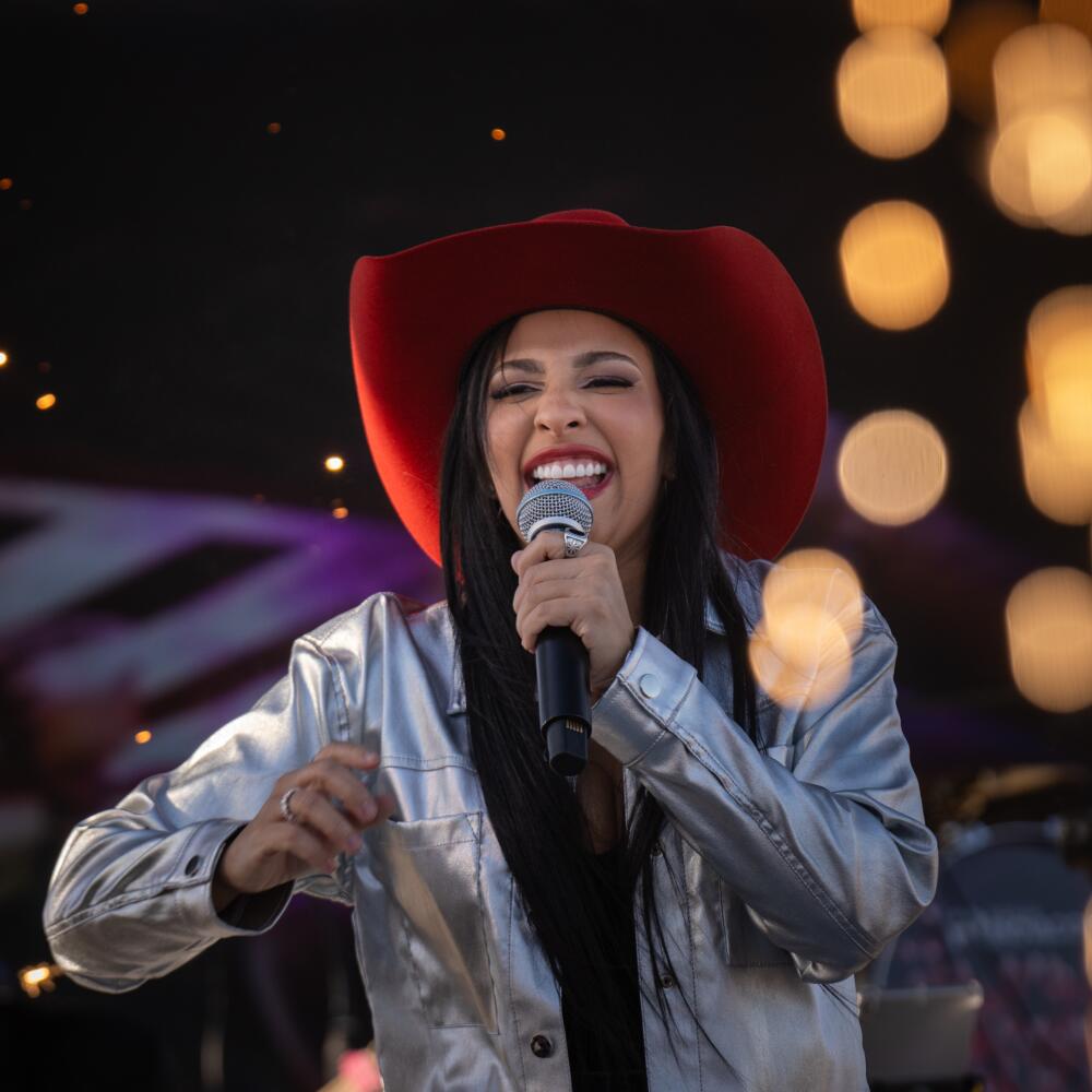  A woman in a silver shirt and burgundy cowboy hat sings into a microphone. 