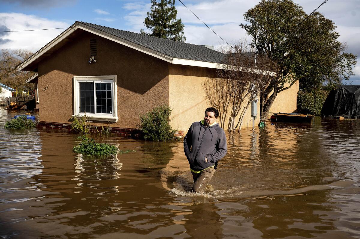 A man in a hoodie trudges through water above his knees. A half-submerged house is in the background.