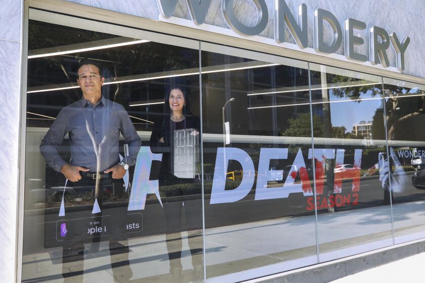 WEST HOLLYWOOD, CA - OCTOBER 27, 2020 - Wondery CEO Hernan Lopez, left, and COO Jen Sargent stand in the display window for the company as podcasts of their programming projects on an LED screen, background, in West Hollywood on October 27, 2020. Lopez is the founder and CEO of Wondery, creates and curates immersive storytelling-based podcasts including Dirty John, Sword & Scale, Hollywood & Crime and Dr. Death, season two. (Genaro Molina / Los Angeles Times)