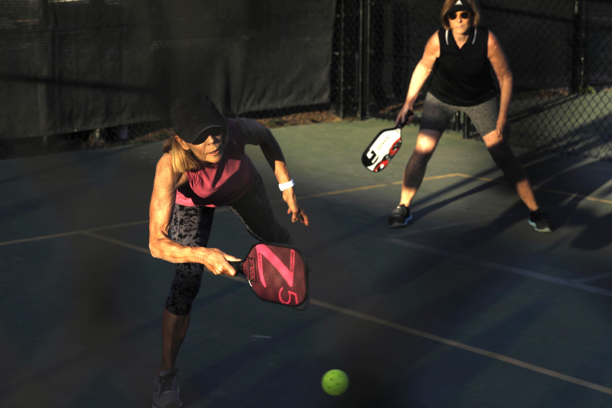 Two people in hats and sunglasses play pickleball on a court