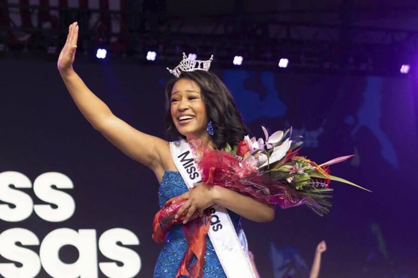 A young Black woman in a glittery blue gown and crown waving her right arm and holding a bouquet