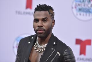 Jason Derulo in a black Moto jacket and large chains posing at a red carpet. 