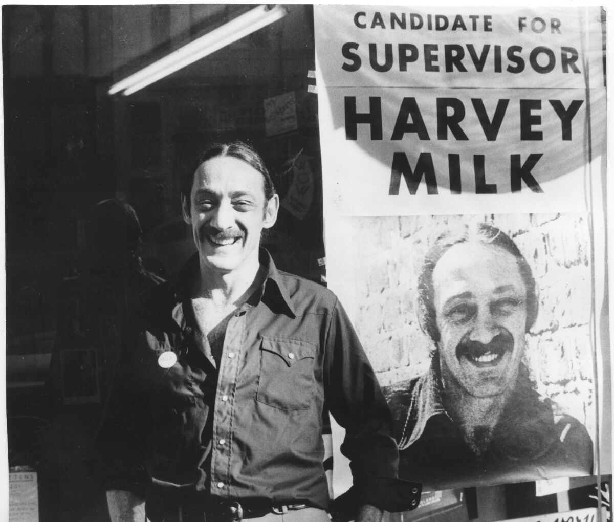 A smiling man in a button-front denim shirt stands next to a poster that reads, "Candidate for supervisor Harvey Milk."