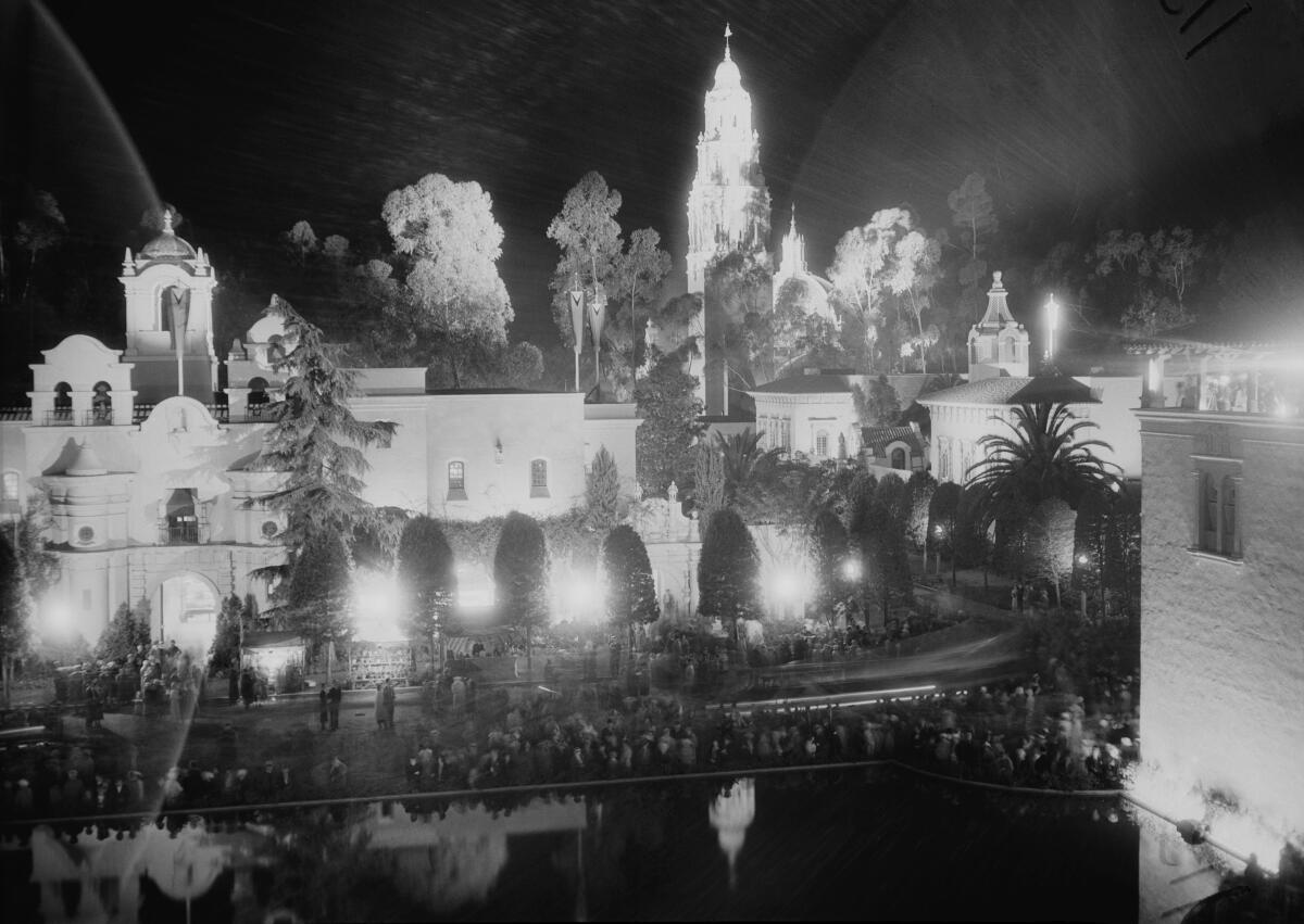 Spotlights illuminate a series of buildings. A crowd fills the street in front. 