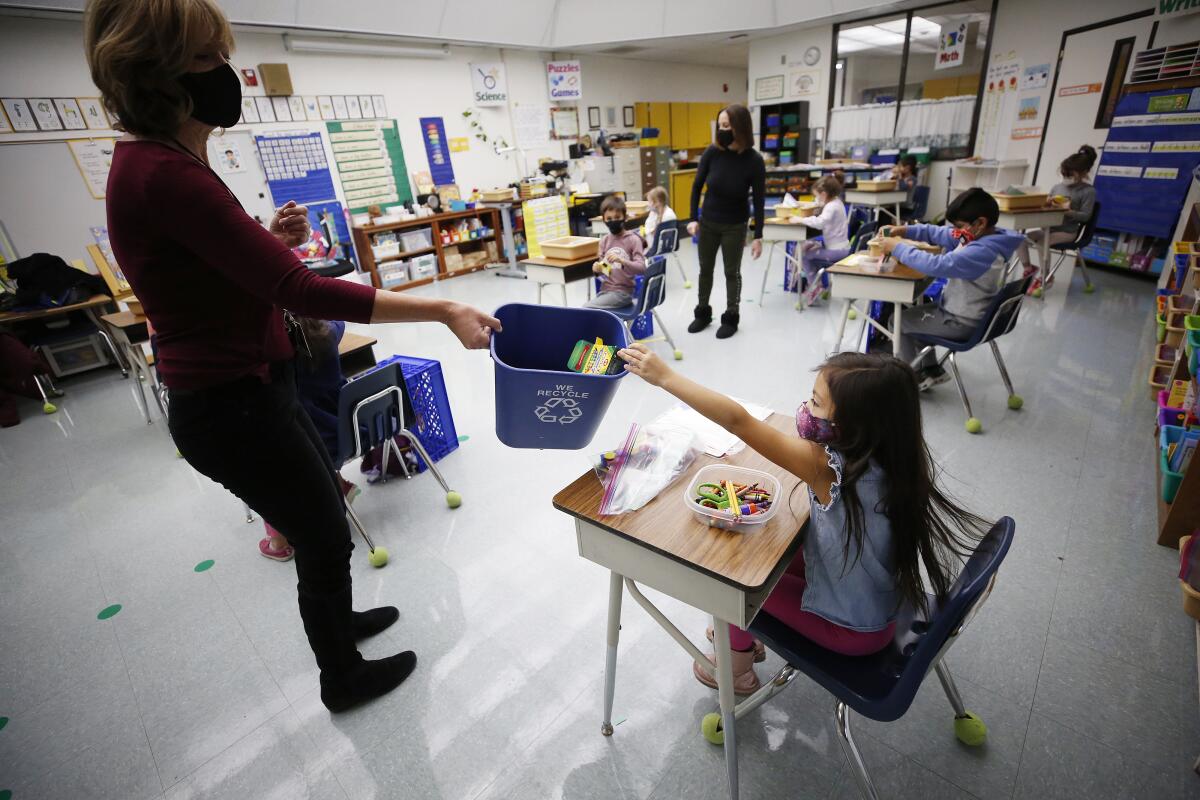 A teacher in a mask holds a recycling bin out to a young girl at a desk