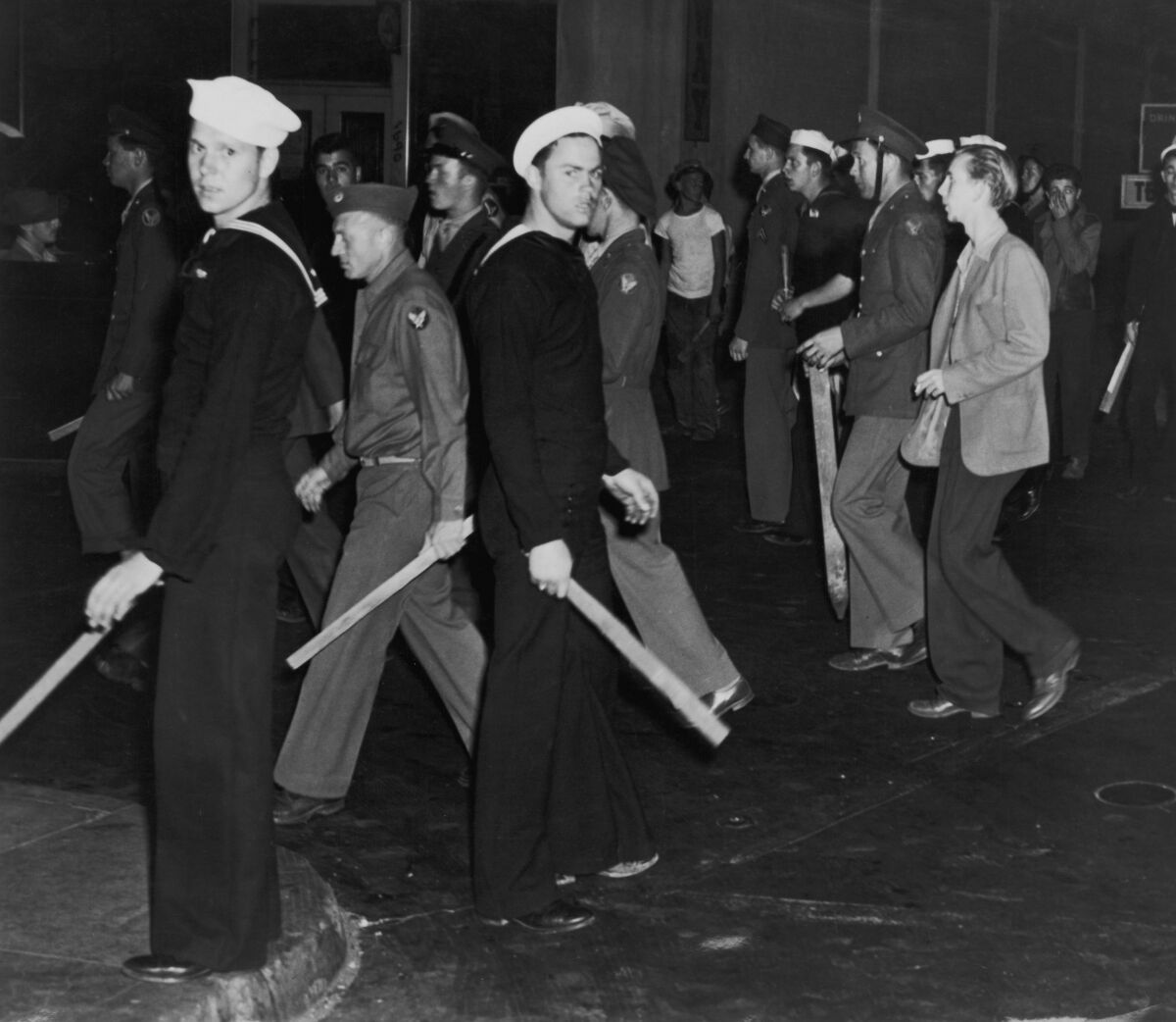 People in sailor and armed forces uniforms walk on a street, some holding sticks.