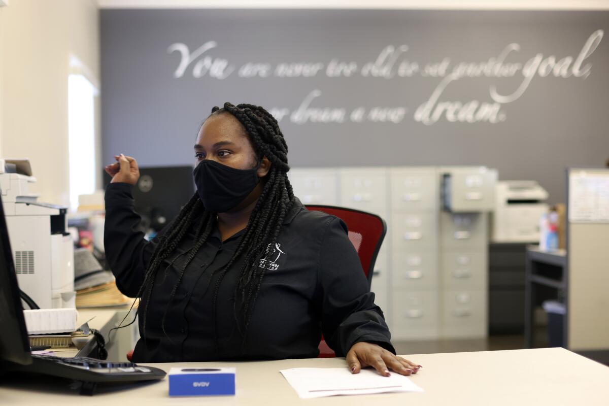 A woman sits at a desk wearing a mask and looking at a computer. 
