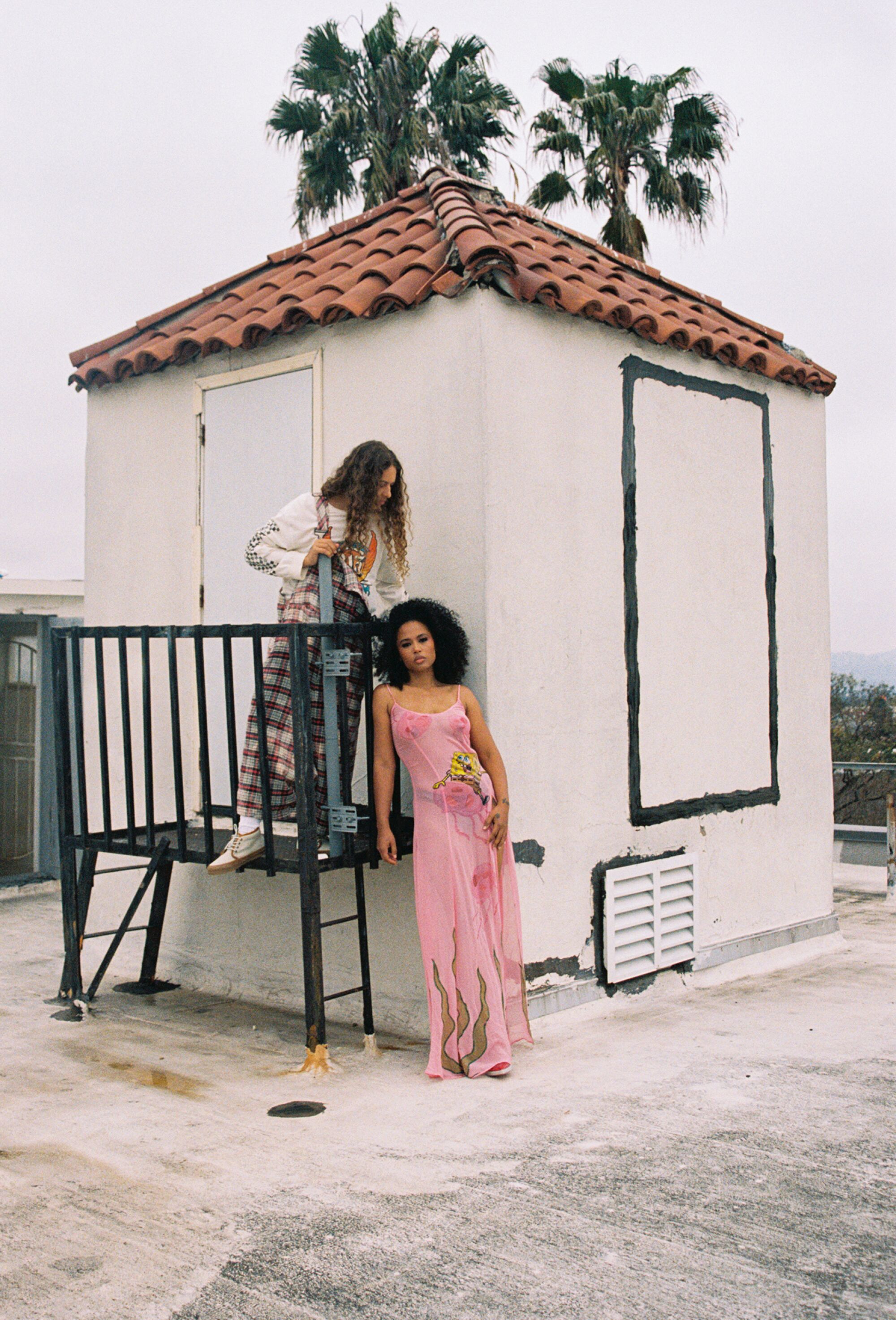 Two people stand near a small stucco structure with red tile roof on a rooftop, with palm trees visible behind.