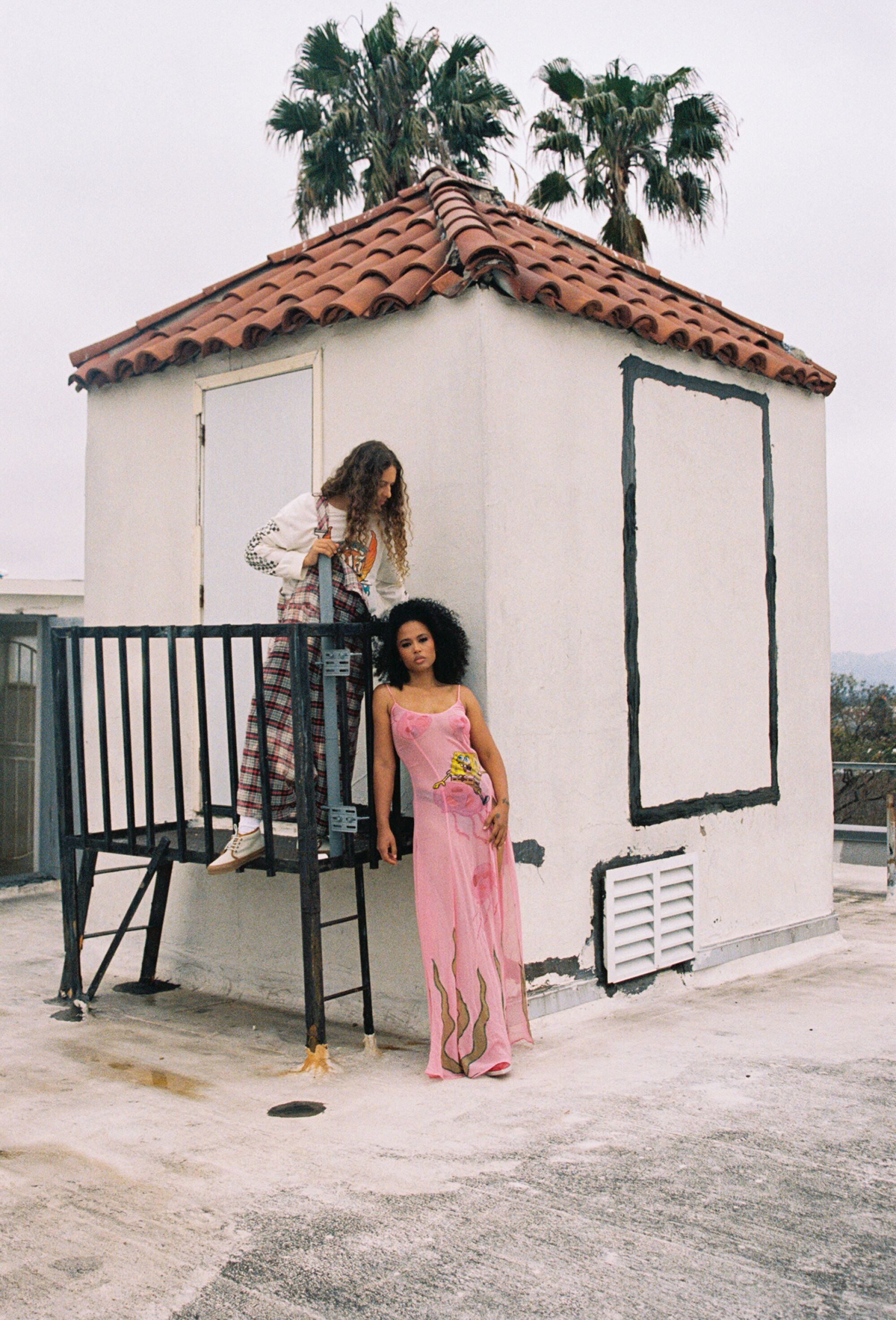 Two people stand near a small stucco structure with red tile roof on a rooftop, with palm trees visible behind. 