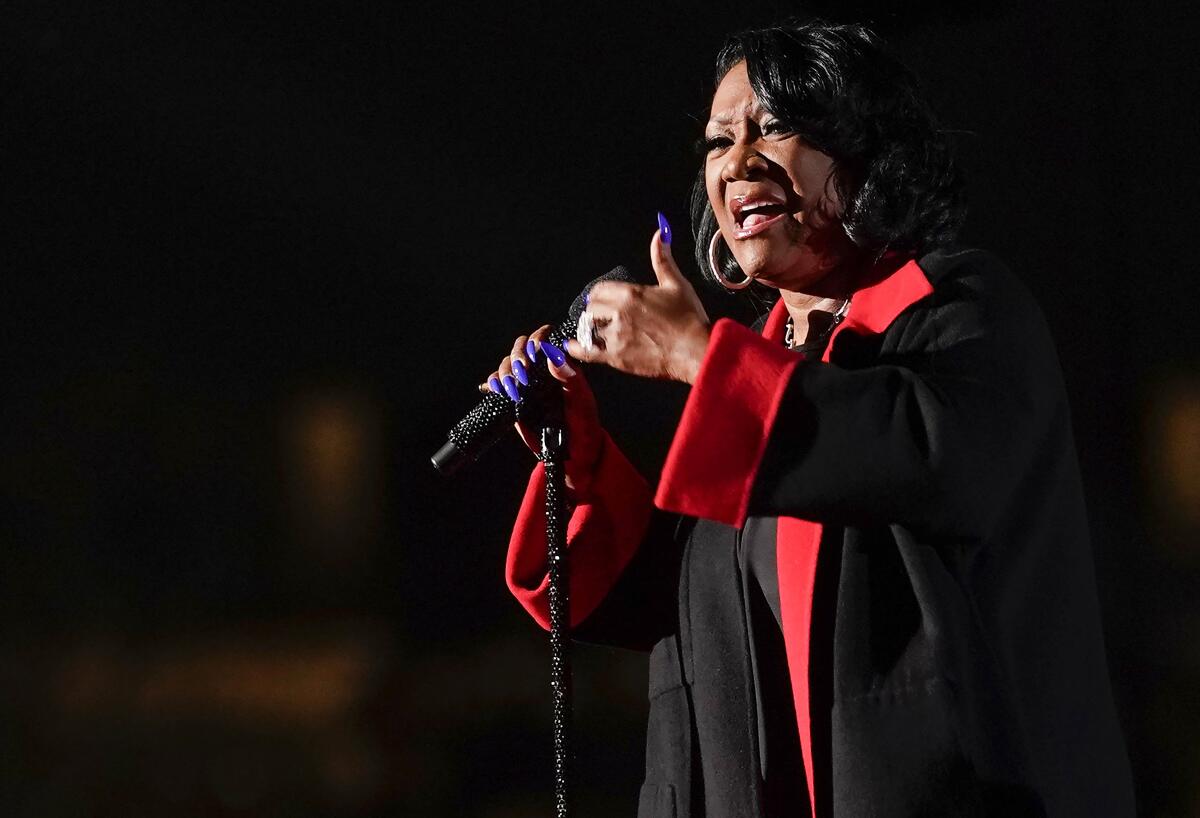 A woman with short black hair wearing a black-and-red coat and singing into a microphone