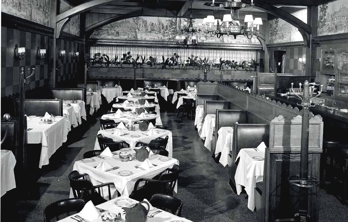 A black-and-white photo of a restaurant interior, with white-tableclothed tables and booths, Gothic wood paneling and beams