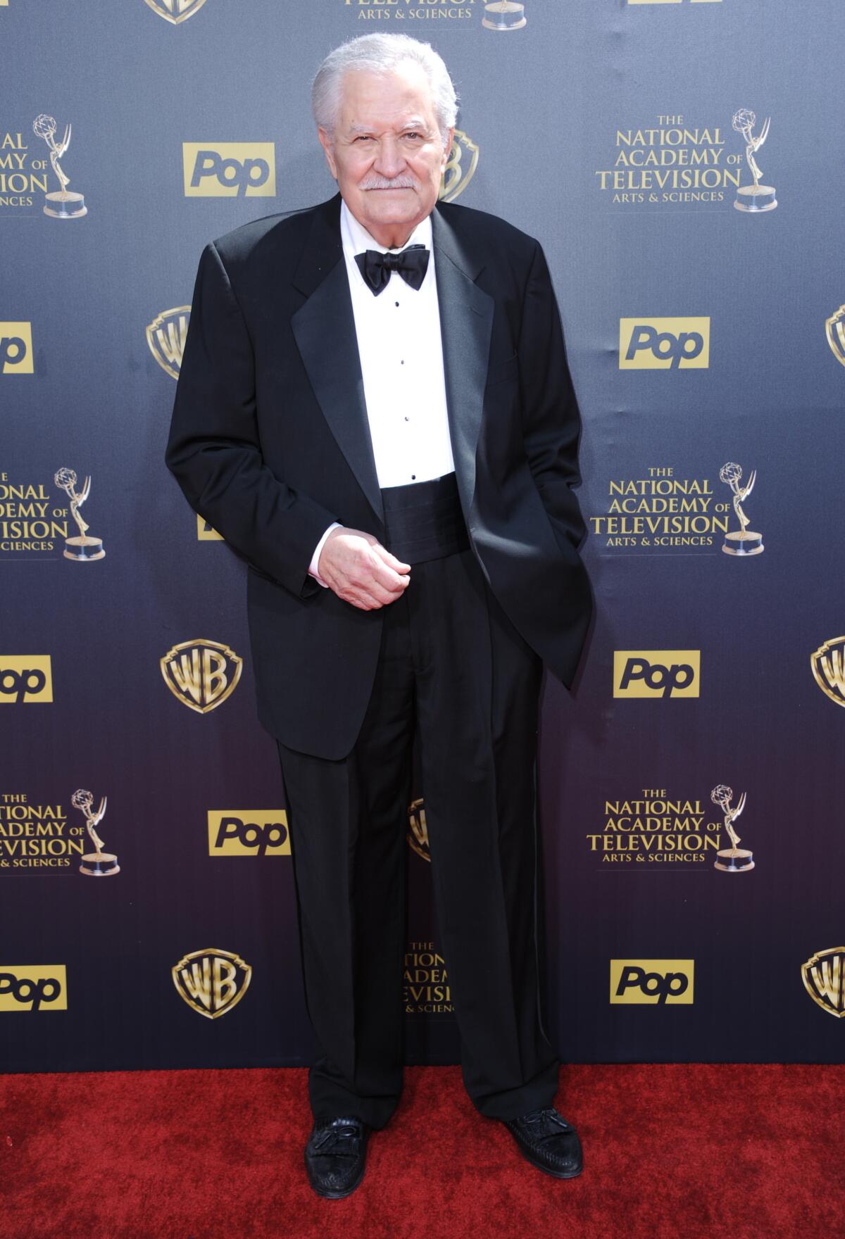 An older man wearing a tuxedo stands in front of a backdrop on a red carpet.