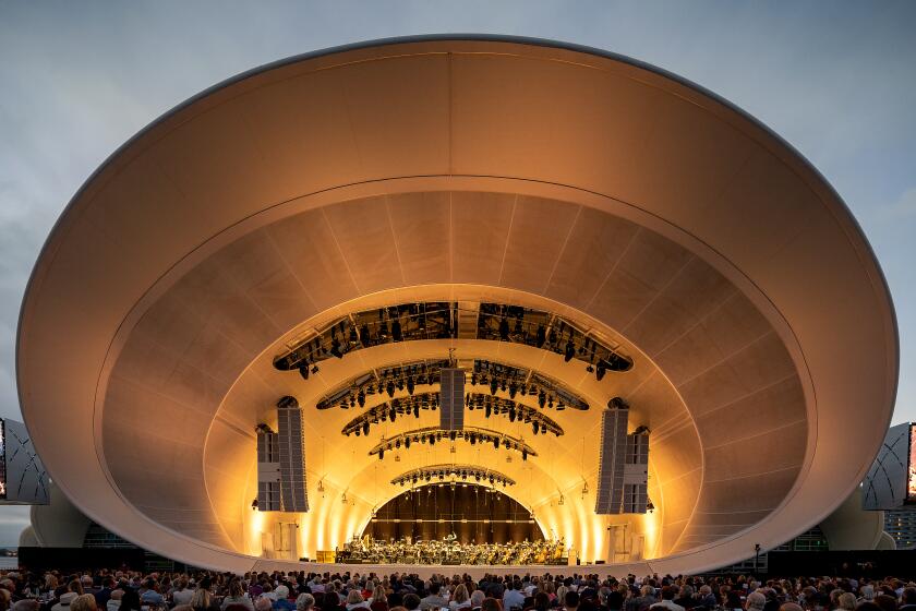 A symphony orchestra performs on the stage of a large white bandshell bathed in warm light.