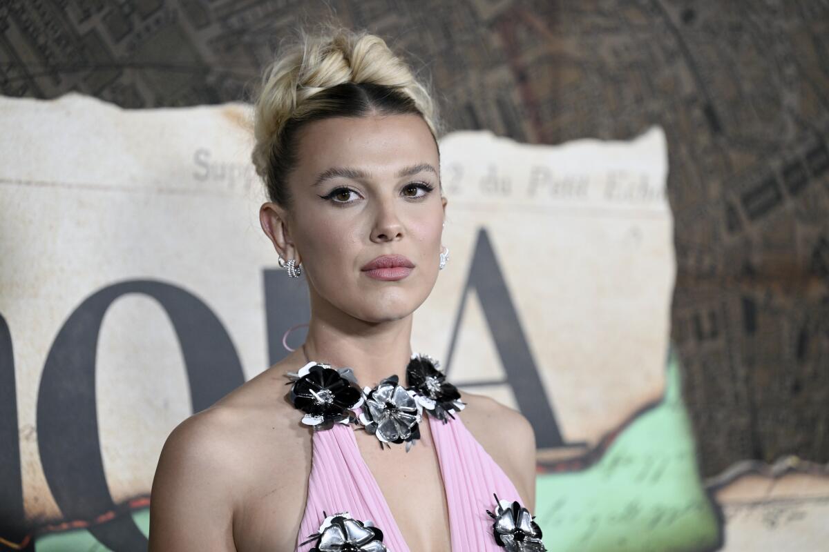 Millie Bobby Brown poses in a pink halter dress with black-and-white flowers,  with her hair up.