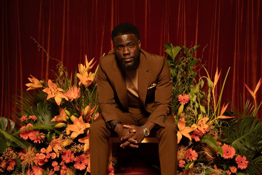 Kevin Hart in a brown suit and shoes sitting among orange flowers and green leaves and a copper carpet
