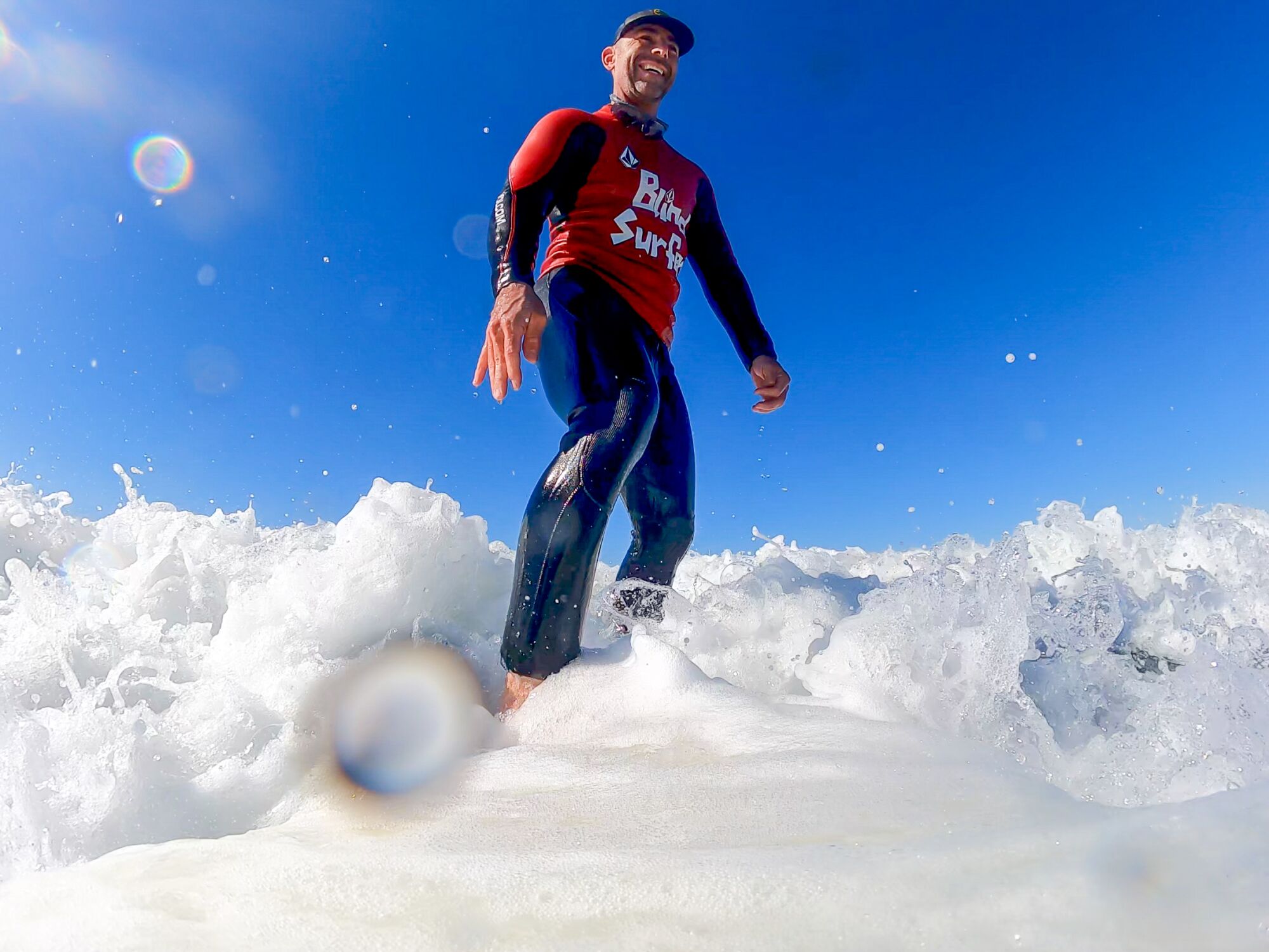 A man  grins while standing on a surfboard in the ocean.