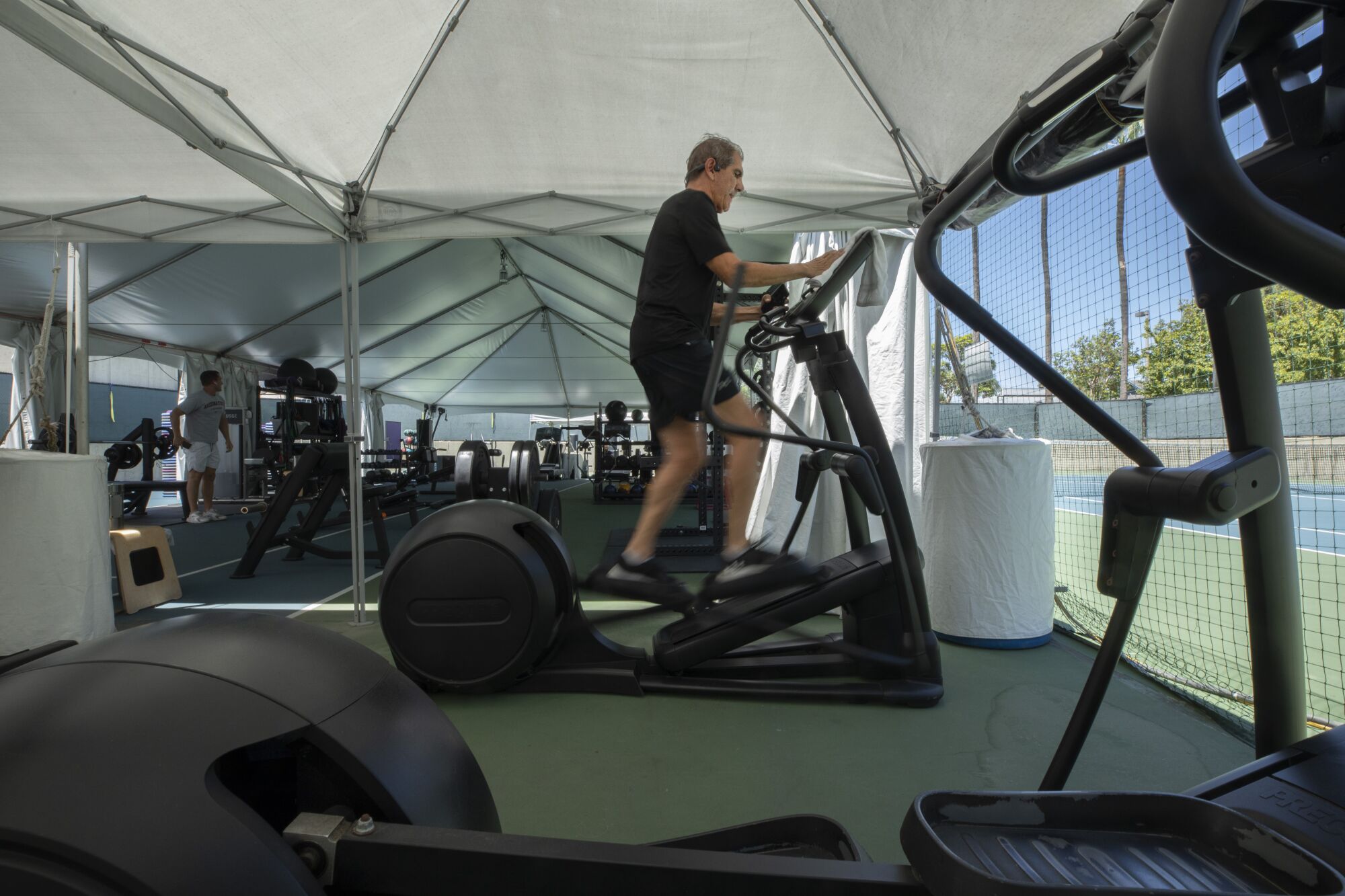 A man works out on an outdoor elliptical machine in an outdoor gym under a tent. 