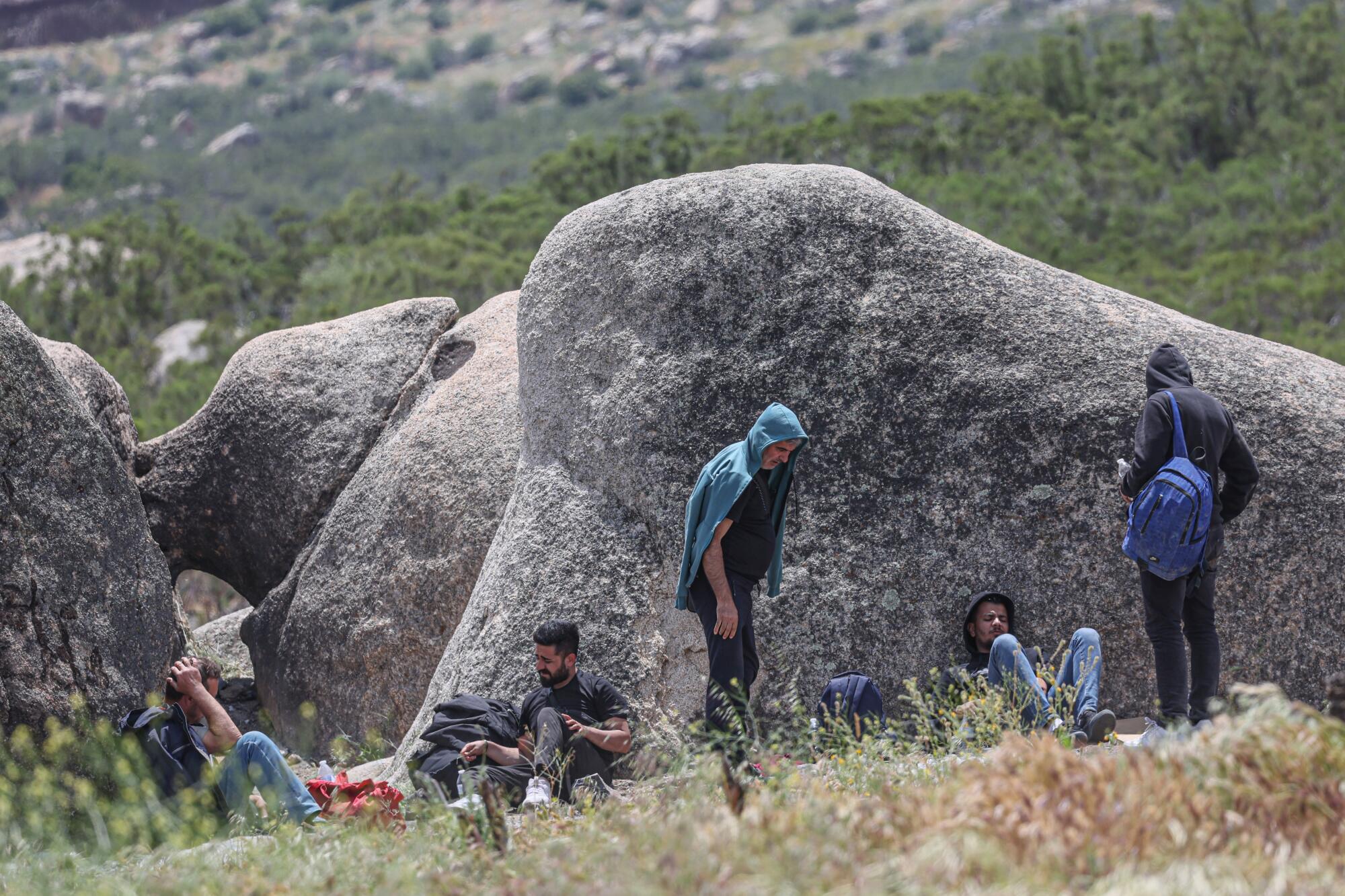 People sit and stand near large rocks. 