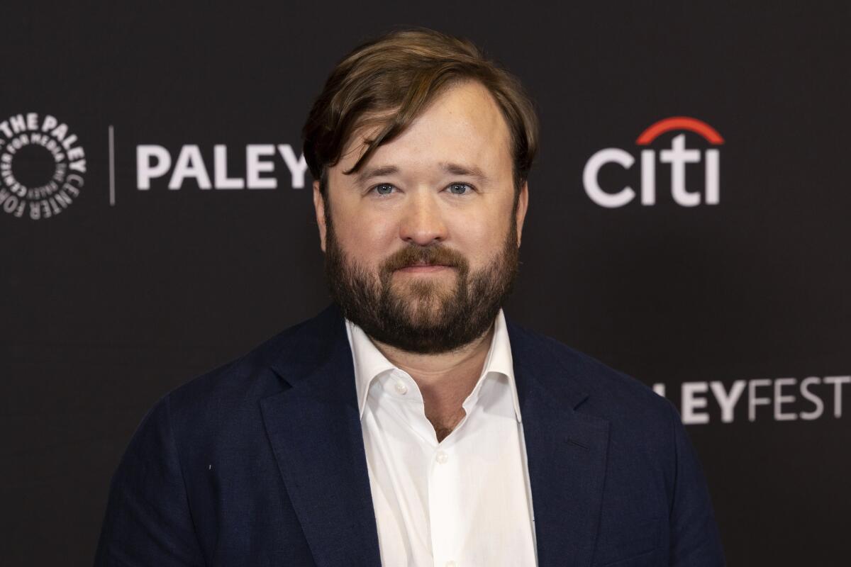 Haley Joel Osment in a dress shirt and blazer posing in front of a black backdrop with white text