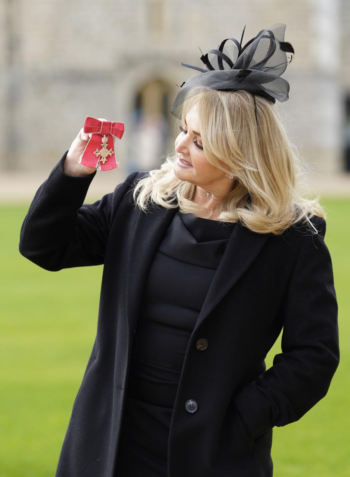 Bonnie Tyler in a black dress and black fascinator holding a small, red piece of fabric with a broach on it.