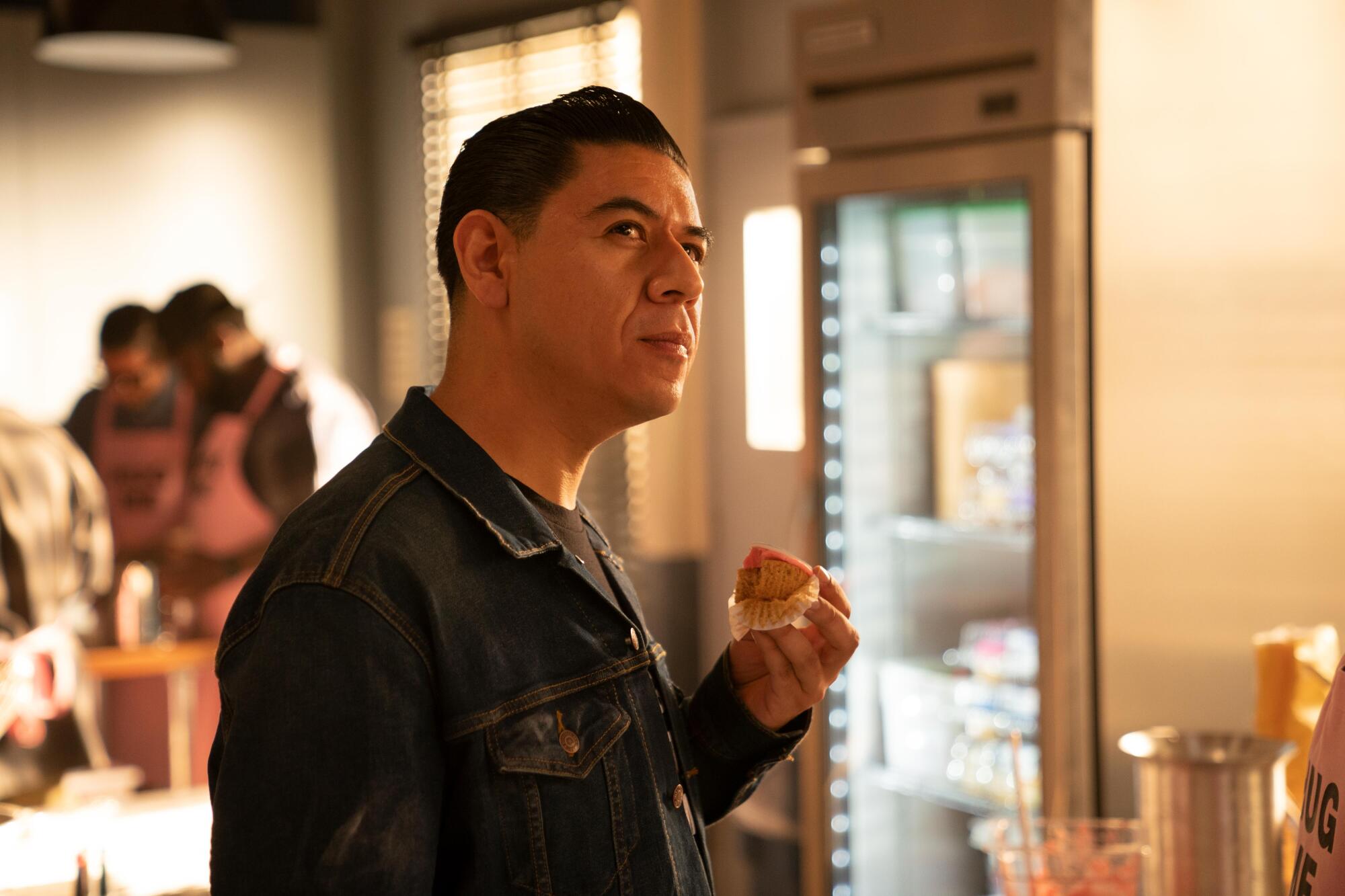 A man in a denim jacket looks contemplative as he eats a cupcake with pink frosting.