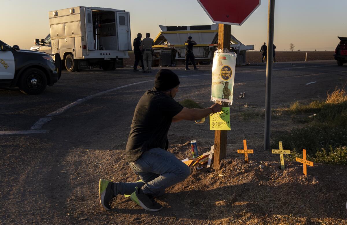 A man tapes a sign to homemade crosses next to a rural road with trucks and police cars behind him