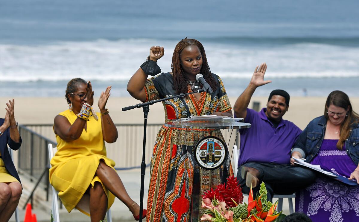 A Black woman stands at a lectern on a beach and speaks, raising her fist