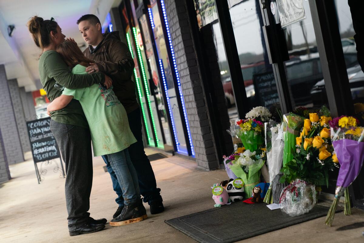People hug outside a storefront with flowers and stuffed animals placed near it