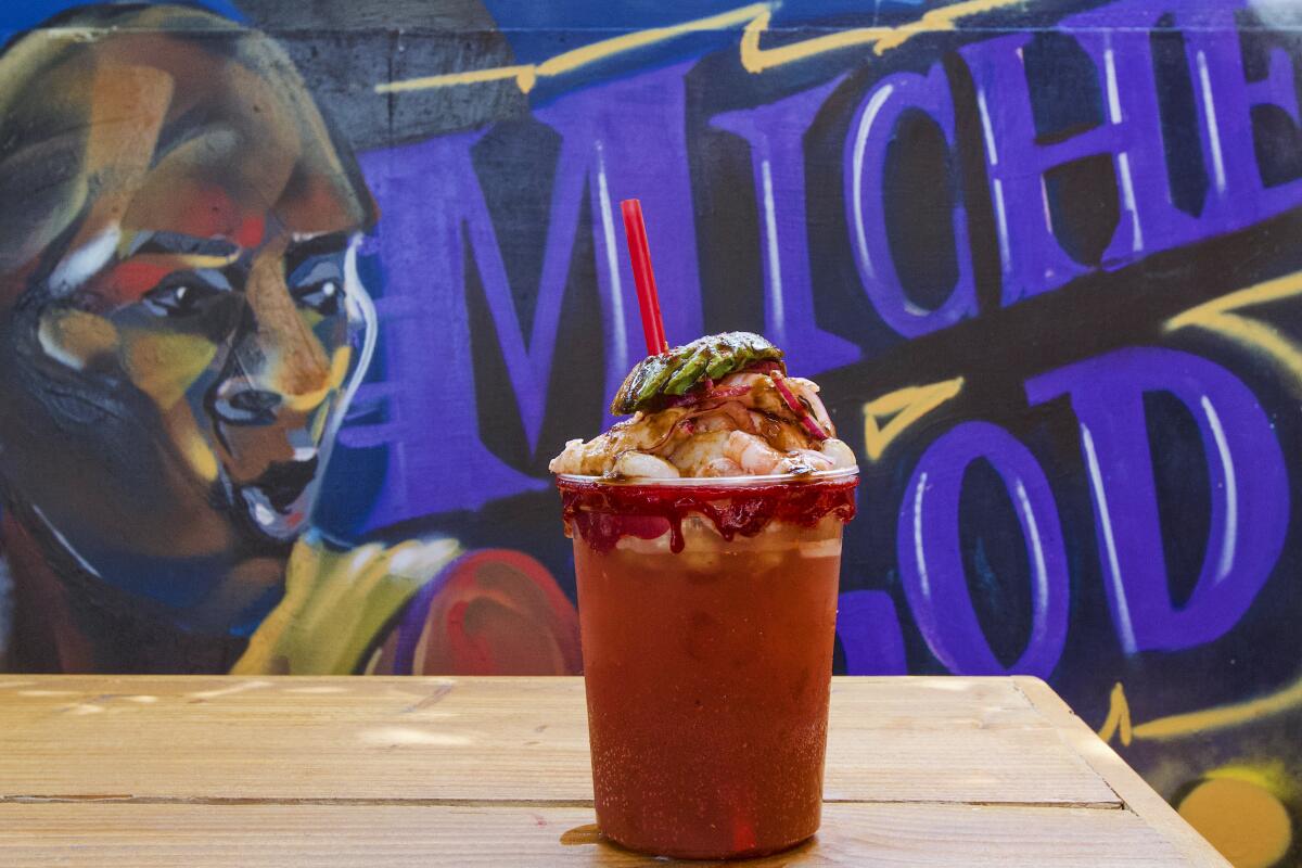 A botana michelada, stacked with shrimp-and-scallop aguachile, in front of a mural with the words Miche God.