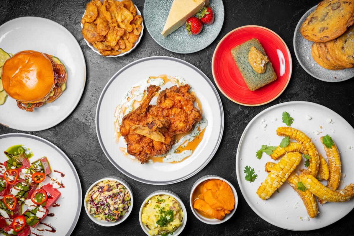 An overhead photo of chicken thigh, chicken sandwich, and sides such as corn ribs, cornbread, cookies and watermelon salad.