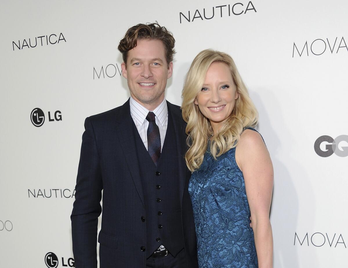 A man wearing a suit and tie and a woman in a blue dress smile in front of a step-and-repeat banner.