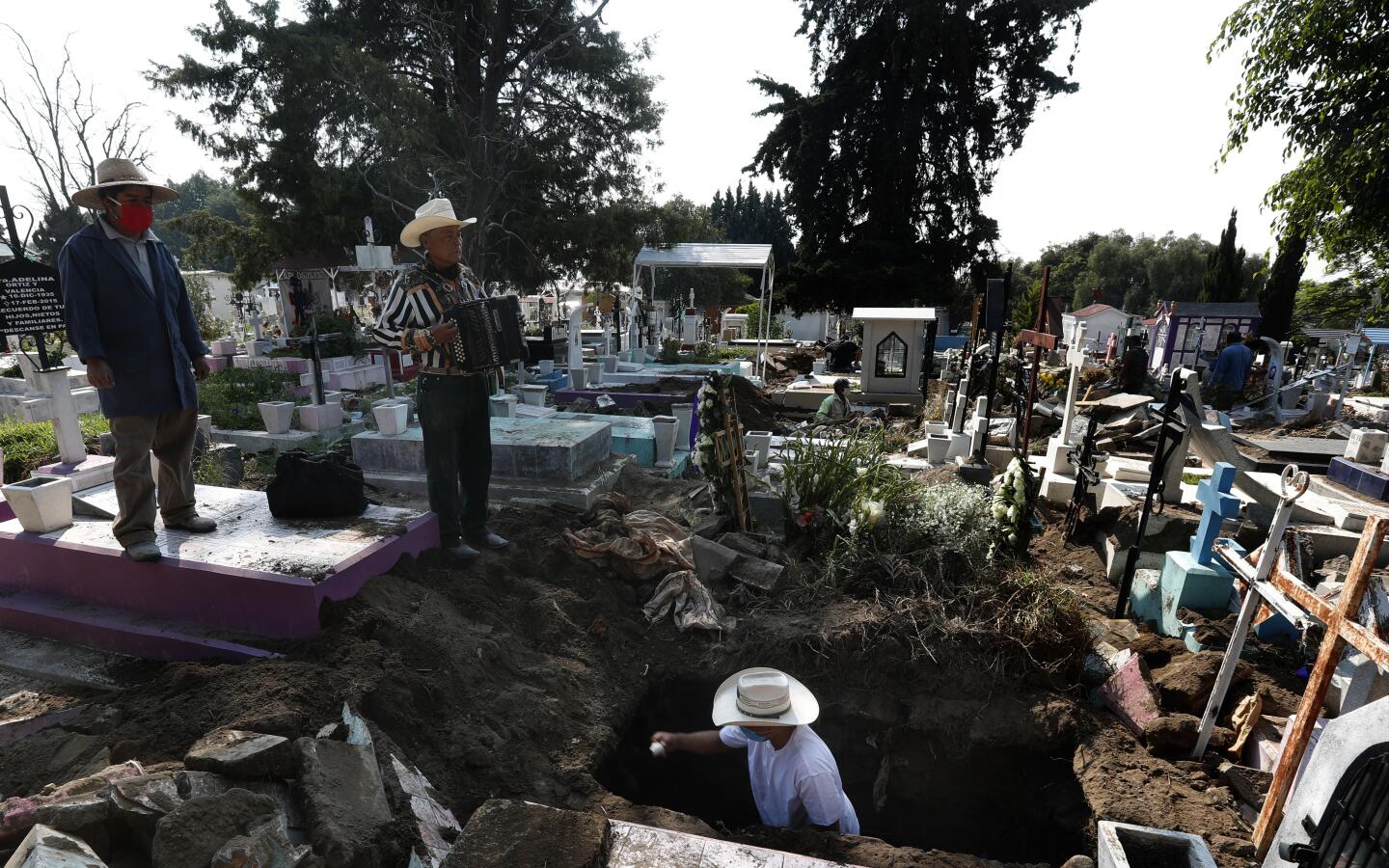 El músico de cementerio Víctor Dzib Cima, de 70 años, toca su acordeón mientras espera clientes cuando trabajadores de cementerio retiran ataúdes de tumbas que pertenecen a familiares que dejaron de pagar alquiler en el cementerio San Nicolás Tolentino, en donde ha trabajado a cambio de propinas durante 22 años en la alcaldía Iztapalapa en Ciudad de México el viernes 22 de mayo de 2020. El cementerio hace espacio para más entierros durante la pandemia de COVID-19 y se acostumbra que los cementerios en México alquilen, en lugar de vender, los sitios de entierro. (AP Foto/Marco Ugarte)