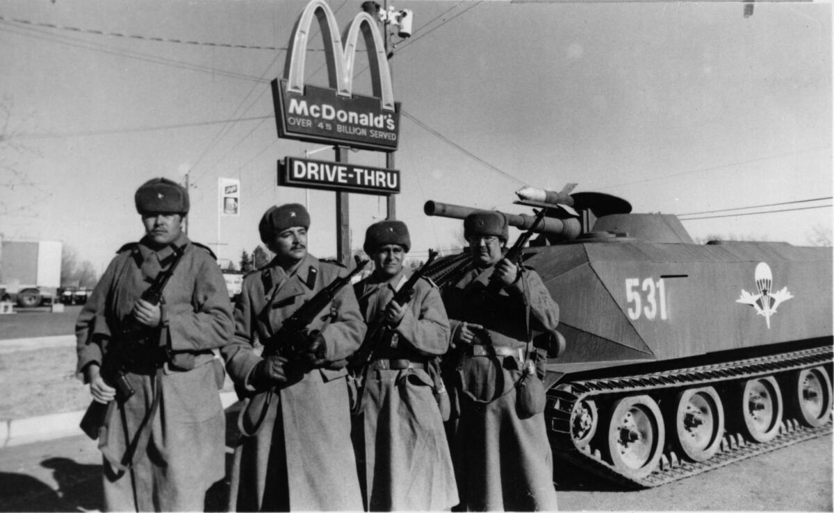 Men in long coats and holding rifles stand next to a tank in front of a McDonald's.