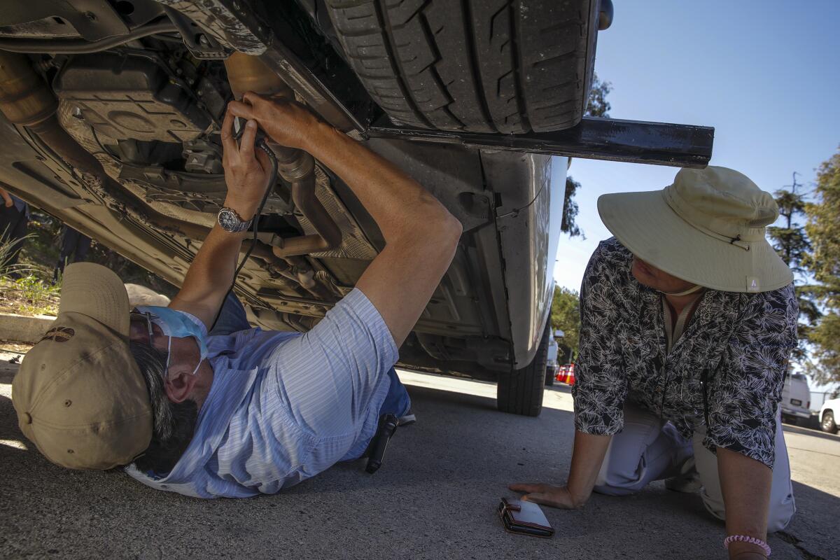 A man in a baseball cap lies on the ground and uses a tool on the underside of a car as a woman kneels nearby and looks on.