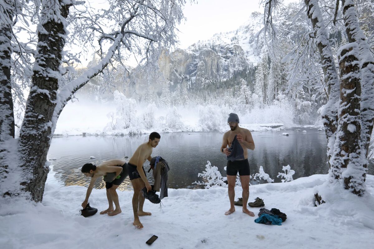 Three men in their underwear gather their clothing on a snowy river bank.
