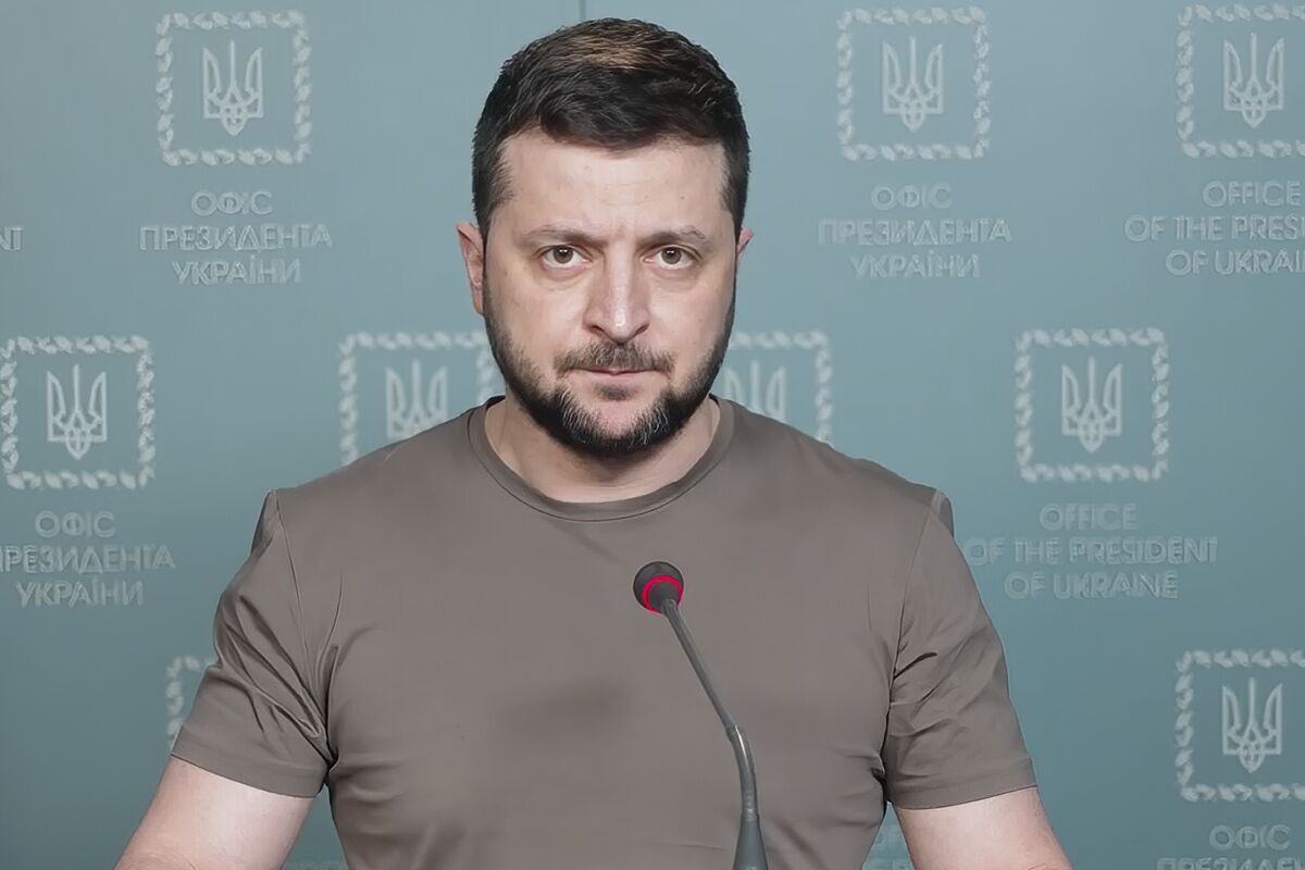A man with dark hair and a beard, wearing a brown T-shirt, speaks before a microphone