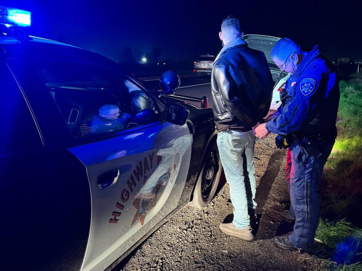 An officer stands behind a man next to a highway patrol vehicle at night.