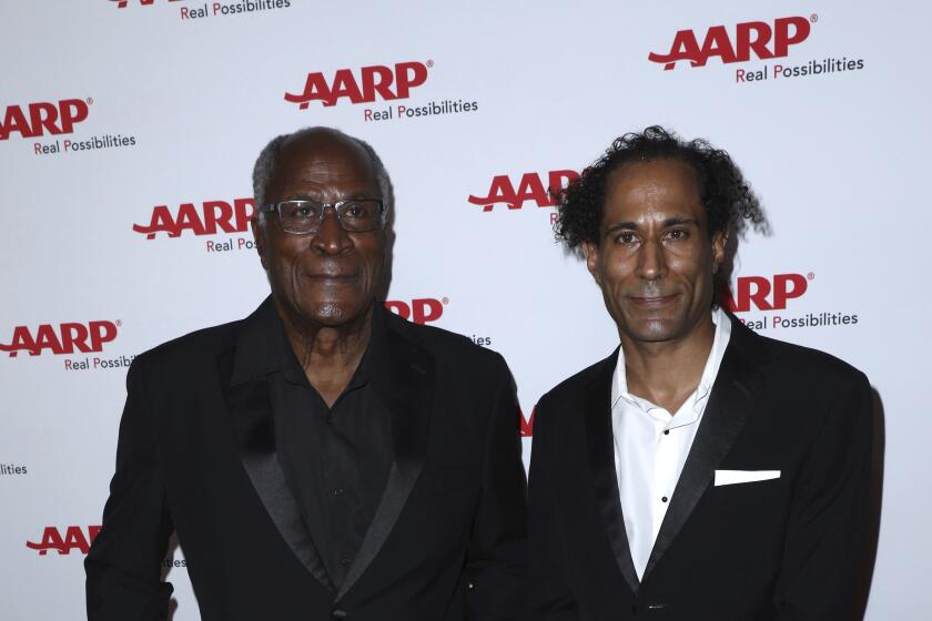 John Amos and K.C. Amos pose together on a red carpet in black formal attire.