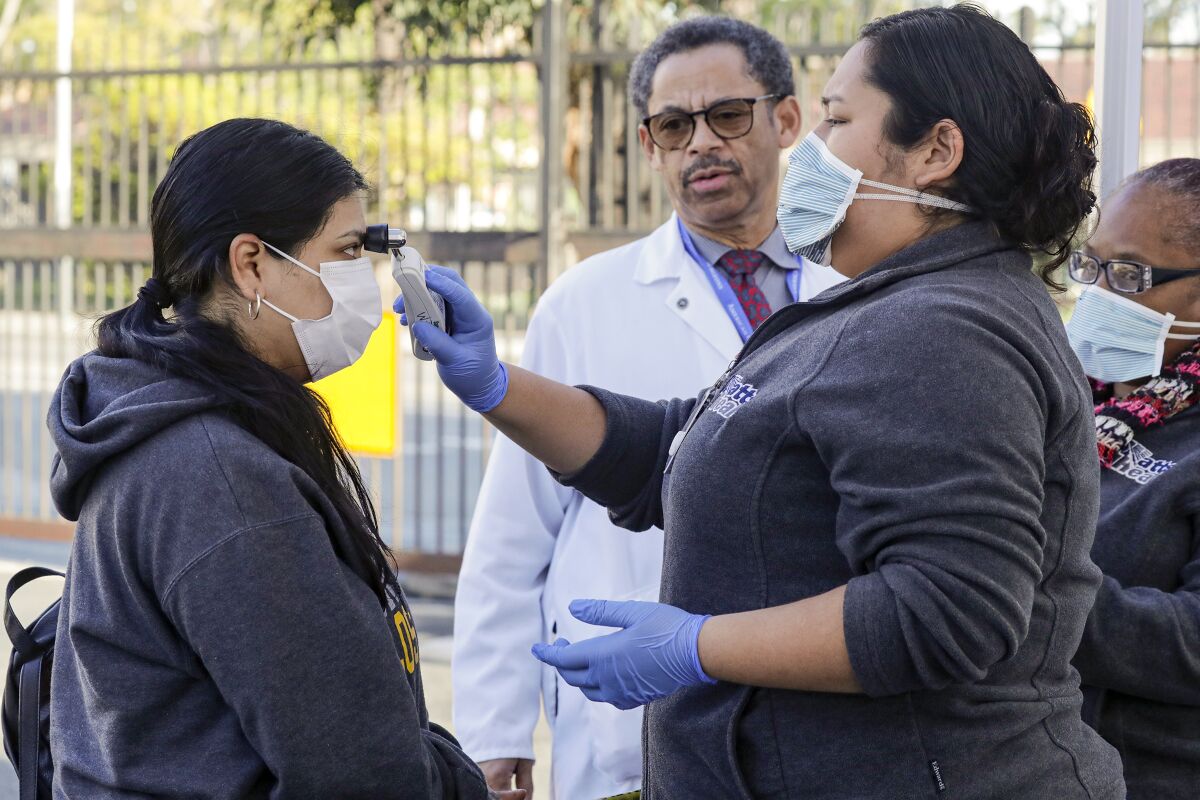 Dr. Oliver Brooks, center, looks on as healthcare worker Lucy Arias, right, checks a patient, at a for COVID-19 screening station, for fever at Watts Health Center, Los Angeles.