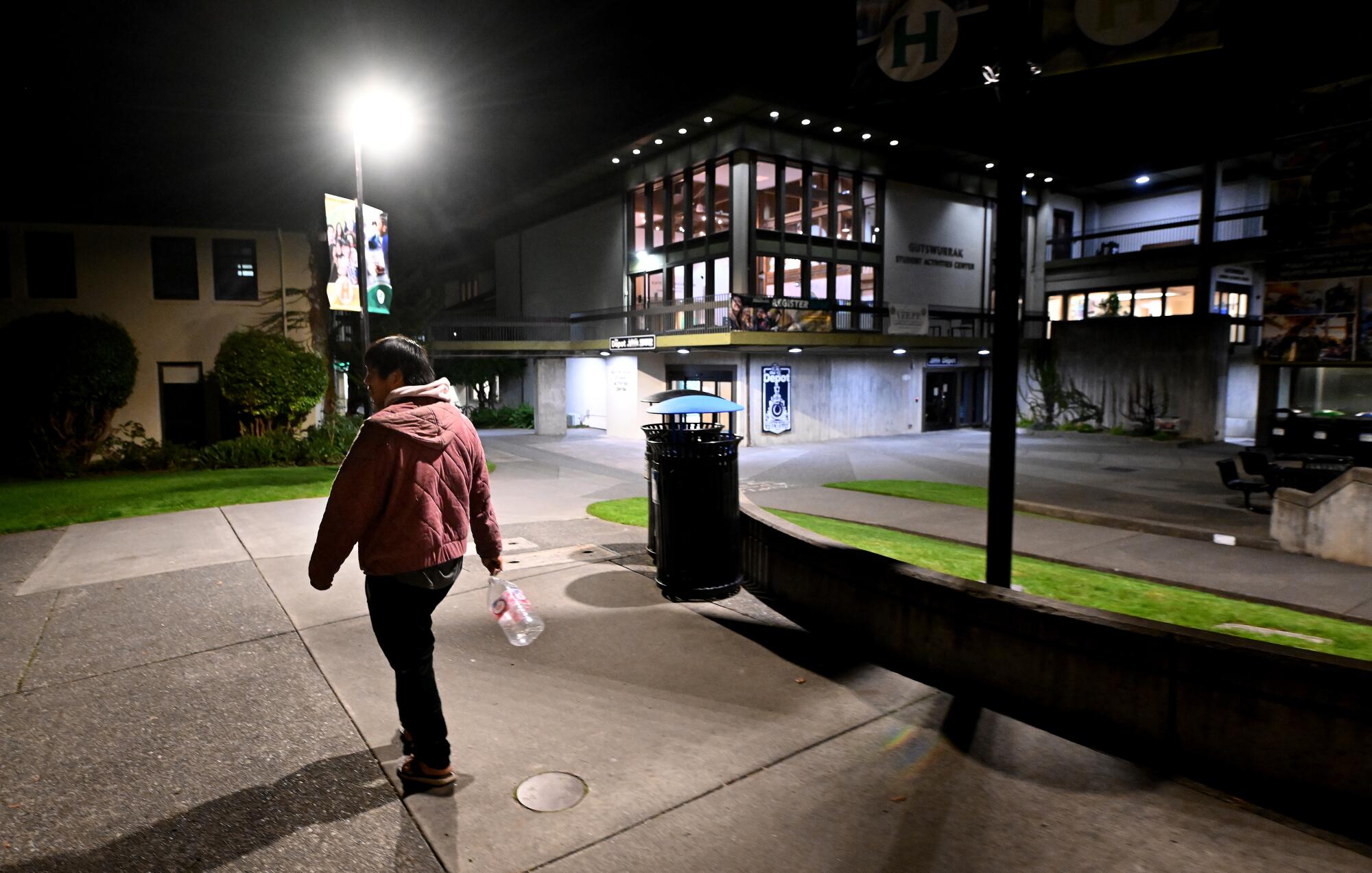 A person carrying an empty water container walks toward buildings at night.